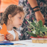 A girl at a Monarch Festival holds a butterfly cutout while examining Monarch caterpillars on milkweed leaves.