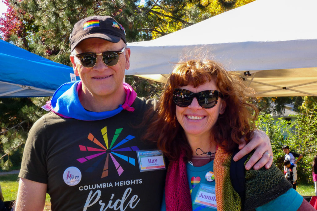 People at a sunny outdoor PRIDE festival, wearing PRIDE apparel and pronoun pins.